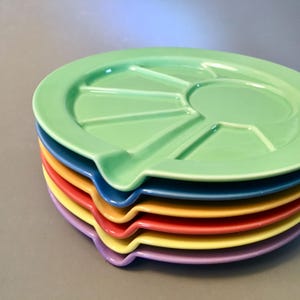May include: A stack of colorful ceramic divided plates. The top plate is light green, with a central circular section and radiating dividers. Below, plates in blue, red, yellow, and purple are visible. Each plate has a small, curved handle.