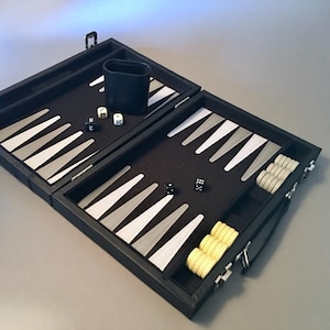 May include: A black backgammon set with white and gray triangles, dice, and light yellow game pieces. The set is open, revealing the playing surface and storage compartments. A black cup is also included.