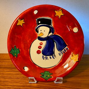 May include: A round, hand-painted ceramic plate with a snowman design. The plate features a red background with a snowman wearing a black top hat and blue scarf. Yellow stars, green trees, and white dots decorate the plate's border. The plate is displayed on a clear stand.