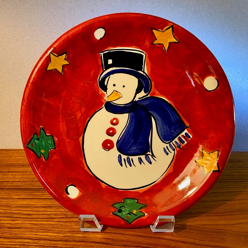May include: A round, hand-painted ceramic plate with a snowman design. The plate features a red background with a snowman wearing a black top hat and blue scarf. Yellow stars, green trees, and white dots decorate the plate's border. The plate is displayed on a clear stand.