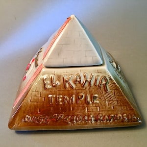 May include: A ceramic pyramid-shaped container with a lid. The pyramid is tan with a white top and features the words "El Kahia Temple" and "Oasis of Cedar Rapids, IA." The container has a decorative design and a pink ribbon.