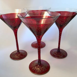 May include: Four red martini glasses with gold swirl designs. The glasses have long stems and round bases, with a gold rim around the top and base. The glasses are set against a white background.