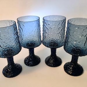 May include: Four blue textured glass goblets with a dark blue base and stem. The glasses have a unique, organic texture on the bowl and base. The goblets are arranged in a row, showcasing their design.