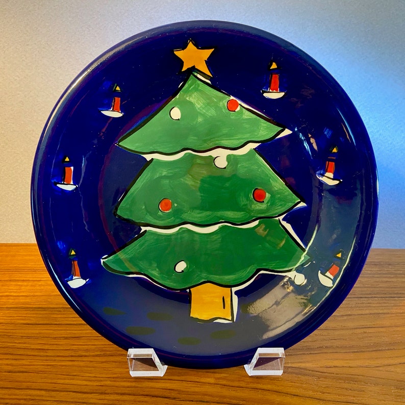 May include: A round, dark blue ceramic plate with a hand-painted Christmas tree design. The green tree has red ornaments and a yellow star. White candles are painted around the rim. The plate is displayed on a clear acrylic stand.