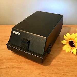 May include: A black Rolodex card file with a rectangular shape and a hinged lid. The word "ROLODEX" is printed on a white label on the front. The file sits on a wooden surface.