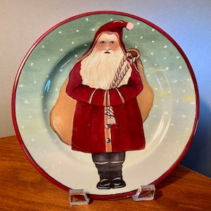 May include: A decorative plate featuring a hand-painted Santa Claus design. Santa is depicted in a red coat and hat, carrying a sack. The plate has a green background with white stars and a red rim, perfect for holiday decor.