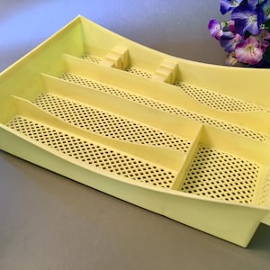 May include: A pale yellow plastic organizer with multiple compartments and a perforated base. The rectangular container has a curved design and is likely used for storing small items. The organizer is empty and sits on a gray surface.