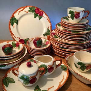 May include: A collection of cream-colored ceramic dinnerware with a red apple and green leaf design. The set includes plates, bowls, cups, saucers, a sugar bowl, a creamer, and a serving platter. The rims are decorated with a brown branch pattern.