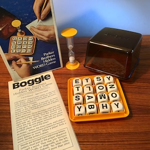 May include: A vintage Boggle word game set. The game includes a yellow cube tray with lettered dice, a timer, and a clear plastic cover. The box and instructions are also visible.