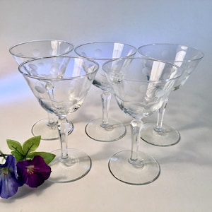 May include: Five clear glass cocktail glasses with etched floral and dot patterns. The glasses have a stemmed base and a wide, shallow bowl. The glasses are arranged on a white surface, with a few purple and blue flowers in the lower left corner.