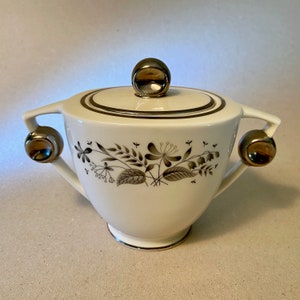 May include: A white ceramic sugar bowl with a lid and two handles. The bowl features a floral design in shades of gray and black. The lid and handles have gold-colored accents. A classic piece for serving sugar.