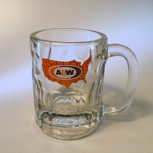 May include: Clear glass mug with a handle, featuring a textured exterior. The mug has an orange and brown graphic of the United States with the A&W logo. The mug is designed for beverages.
