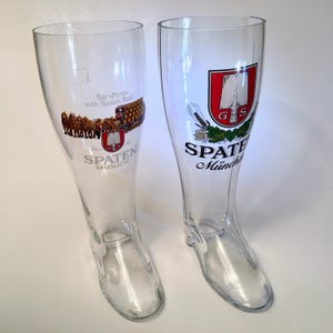 May include: Two clear glass beer boot-shaped mugs. One mug has a Spaten Munich logo with a horse-drawn carriage and the text "Say 'Prost' with Spaten Beer!". The other mug has a Spaten Munich logo with a red shield.