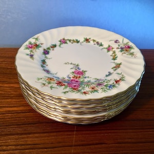 May include: A stack of white porcelain plates with a floral design and gold rims. The plates have a scalloped edge and are decorated with a garland of pink, purple, and yellow flowers.