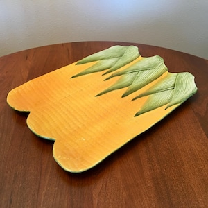 May include: A rectangular, yellow ceramic serving platter with a scalloped edge. The platter features green corn husk designs at the top. The platter is on a dark wooden table.