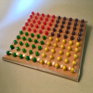 May include: A wooden pegboard game with a grid of holes filled with colorful pegs. The pegs are arranged in rows and columns, with sections of red, green, yellow, and purple pegs. The game is sealed in clear plastic.