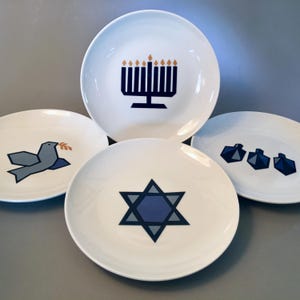 May include: Four white ceramic plates with blue and gold geometric designs. One plate features a menorah, another a dove with an olive branch, a third a Star of David, and the last, four dreidels. The plates are arranged on a gray surface.
