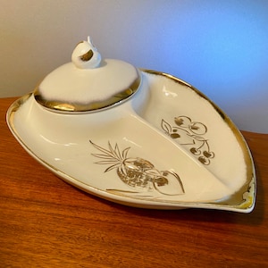 May include: A vintage, white divided serving dish with a lid, featuring gold trim and fruit illustrations. The dish has a leaf-like shape with two compartments. The lid has a small, decorative handle. The dish is likely made of ceramic or porcelain.
