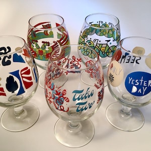 May include: Five clear glass goblets with varied colorful graphic designs. One features red and green rooster art, another a green bicycle, one has red and blue abstract shapes, one displays red floral patterns with the text Take Two, and one has blue circles with the text YESTER DAY.