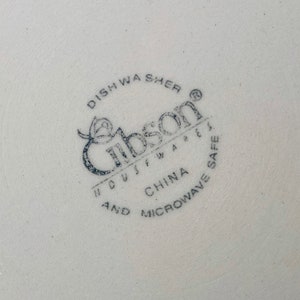 May include: Close-up of a white ceramic plate with a blue stamp. The stamp reads "Gibson Housewares" and includes text indicating it is dishwasher and microwave safe, made in China.
