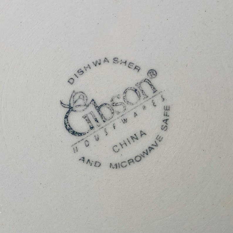 May include: Close-up of a white ceramic plate with a blue stamp. The stamp reads "Gibson Housewares" and includes text indicating it is dishwasher and microwave safe, made in China.