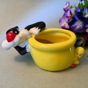 May include: A yellow ceramic planter featuring Sylvester the Cat and Tweety Bird. Sylvester is perched on the side, with Tweety peeking from the other side. The planter has a round shape and is painted in bright colors.