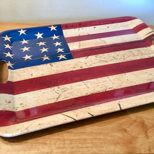 May include: Rectangular serving tray with a distressed American flag design. The tray features a blue corner with white stars and red and white stripes. It has two oval cut-out handles.