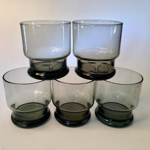 May include: Five clear, gray-tinted drinking glasses. Each glass has a cylindrical body and a dark green, rounded base. The glasses are stacked, showcasing their design and color variations. Ideal for serving beverages.