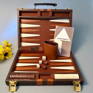 May include: A brown leather backgammon set in a briefcase-style case with a felt interior featuring brown and white triangular points. The set includes a brown leather dice cup, two white dice with numbers, two brown dice with dots, and brown and white checkers.