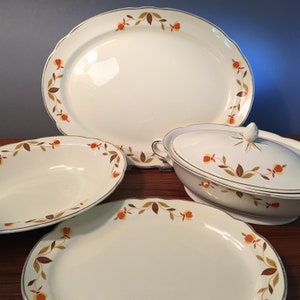 May include: A vintage white ceramic dinnerware set featuring an oval platter, two serving dishes, and a covered serving bowl. Each piece is adorned with a decorative border of brown and yellow leaves and red flowers, with gold trim.