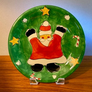 May include: A round, hand-painted Christmas plate with a green background. The plate features a cartoon Santa Claus in a red suit with black boots and a white beard. Yellow stars and candy canes decorate the plate's border. The plate is displayed on a clear stand.