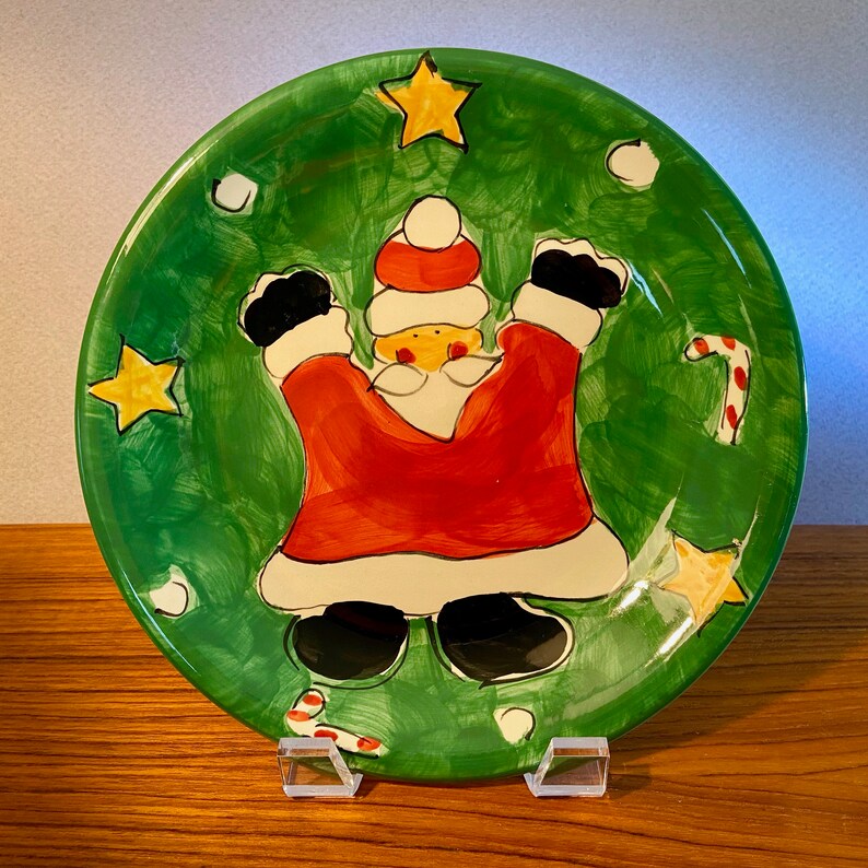 May include: A round, hand-painted Christmas plate with a green background. The plate features a cartoon Santa Claus in a red suit with black boots and a white beard. Yellow stars and candy canes decorate the plate's border. The plate is displayed on a clear stand.