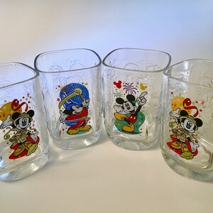 May include: Four clear square-shaped drinking glasses with embossed designs. Each glass features a different colorful image of Mickey Mouse in celebratory poses. The glasses have a curved base with the year "2000" printed on them.