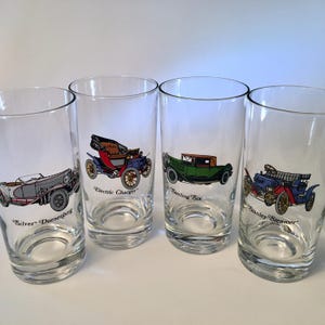 May include: Set of four clear drinking glasses featuring vintage car illustrations. Each glass displays a different car: Silver Duesenberg, Electric Charger, Sterling Six, and Stanley Steamer. The cars are in various colors, including red, green, and blue.
