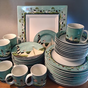 May include: A collection of coastal-themed dinnerware featuring a lighthouse, sailboats, and seaside houses. The set includes plates, mugs, and a large square serving platter. The color palette is primarily blue, white, and green, with a nautical design.