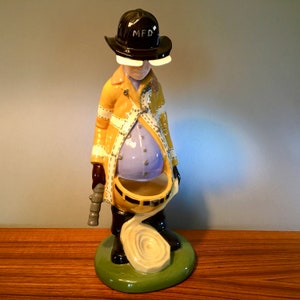 May include: A ceramic figurine of a firefighter in a yellow coat with white stripes and a black helmet marked "MFD". The figure has a blue belly and holds a hose and a rolled-up hose on a green base.