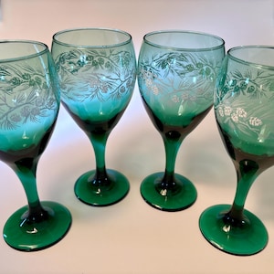 May include: Four green wine glasses with a silver pine needle and pine cone design. The glasses have a dark green stem and base, and a clear bowl. The glasses are arranged in a row.