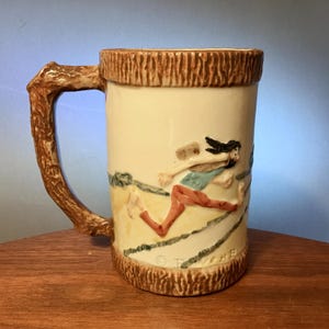 May include: Cream-colored ceramic mug with a brown textured rim and handle. The mug features a relief design of a running figure in red pants and a blue shirt. The handle is shaped like a tree branch.