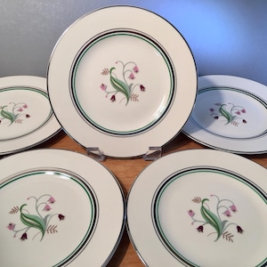 May include: Set of five white ceramic plates with a floral design. Each plate features a central image of pink bell-shaped flowers, green leaves, and brown accents. The plates have a silver rim and a green and brown border.