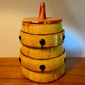 May include: A vintage ceramic cookie jar in the shape of a barrel. The jar is yellow with a wood grain texture and dark brown accents. It has a lid and a brown handle. Black decorative accents are visible.
