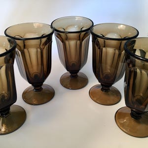 May include: Set of five vintage brown glass goblets. Each goblet features a round base, a stem, and a faceted, flared bowl. The glasses are a warm brown color and appear to be made of pressed glass. The goblets are arranged on a white surface.