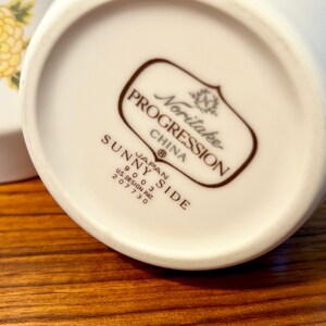 K&ouml;nnte beinhalten: Nahaufnahme des Bodens einer wei&szlig;en Keramik-Tasse mit dem Text "SUNNY SIDE" und "Noritake PROGRESSION CHINA" in Braun. Die Tasse steht auf einer Holzoberfl&auml;che.