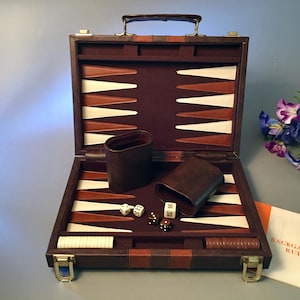 May include: A brown leather-style backgammon set in a briefcase case with a dark felt interior. The board features alternating white and brown triangular points. Included are two dark brown dice cups, white and brown checkers, and four dice.