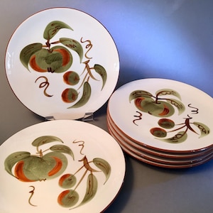 May include: White ceramic plates with a hand-painted apple design in green, orange, and brown. The plates have a brown rim and are arranged in a stack and individually. The apple design includes leaves and curly accents.
