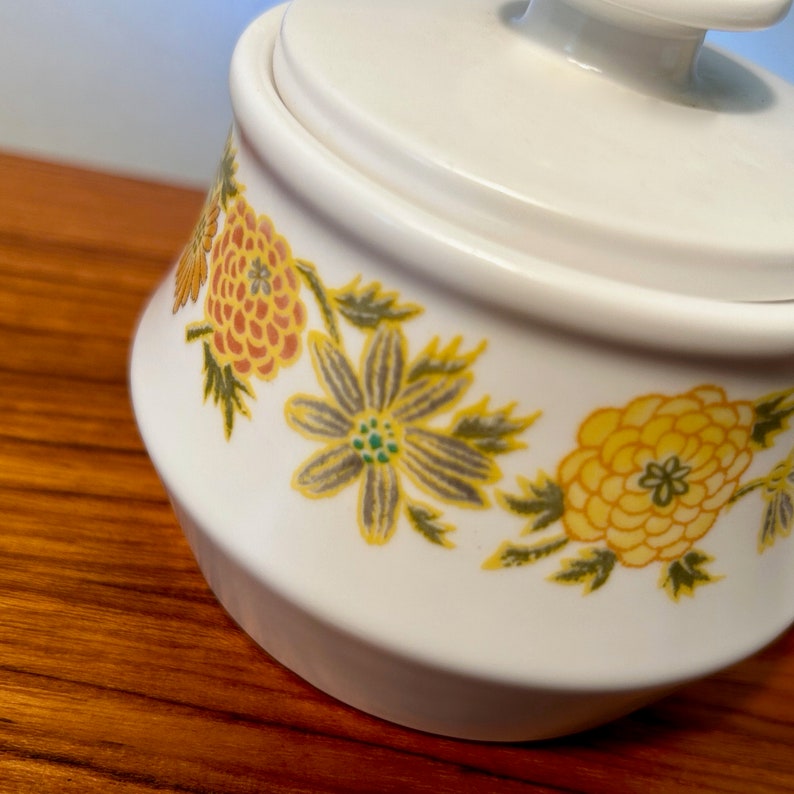 K&ouml;nnte beinhalten: Wei&szlig;er Keramik-Zuckertopf mit Deckel und kleinem Griff. Die Schale ist mit einem floralen Muster in Gelb-, Orange- und Gr&uuml;nt&ouml;nen verziert. Das Design zeigt stilisierte Blumen und Bl&auml;tter.