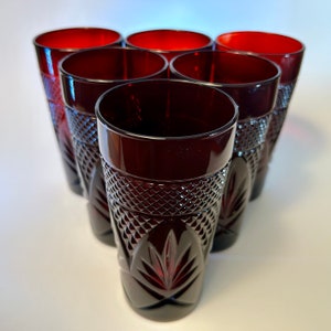 May include: Set of seven ruby red drinking glasses. Each glass has a diamond-cut pattern on the lower half and a smooth rim. The glasses are arranged in a triangular formation, showcasing their elegant design and color.