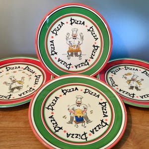 May include: Set of four ceramic pizza plates. Each plate features a chef illustration in the center, surrounded by the word "Pizza" in a circular pattern. The plates have a red and green border design.