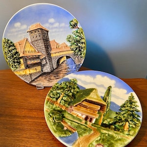 May include: Two decorative ceramic plates with raised relief scenes. One plate depicts a bridge and buildings, the other a house and path. The plates feature blue, green, brown, and yellow hues.