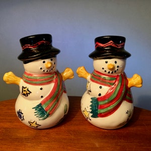 May include: Two ceramic snowman figurines with black top hats, carrot noses, and red, green, and white striped scarves. The snowmen have yellow arms and are decorated with painted stars and other festive details. They are displayed on a wooden surface.