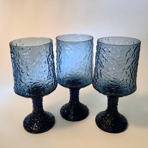 May include: Three blue textured glass goblets with a dark blue base and stem. The goblets have a unique, organic texture on the bowl and base. The glasses are likely for serving beverages.